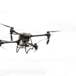 Een commerciële drone vliegt hoog boven een landbouwgebied, met een helder blauwe lucht op de achtergrond.